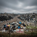 Large illegal dumping site with piles of waste, chemical drums, and discarded electronics near an urban area.